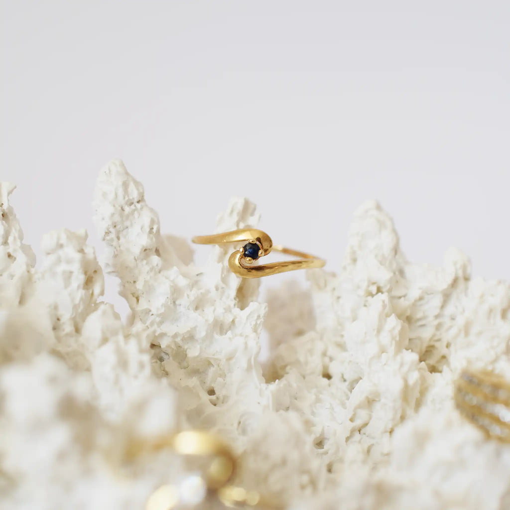 A close-up of a delicate 18K yellow gold ring featuring a single, small, round blue sapphire. The ring's polished gold band has an elegant twisted or bypass design that gracefully cradles the gemstone in its center. The ring is displayed on a textured, white, coral-like stand against a plain light background, highlighting its design and the vibrant blue of the sapphire.