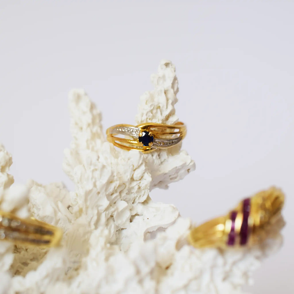 A close-up of an elegant 18K two-tone gold ring, prominently featuring a vibrant blue sapphire set in its center. The ring's design consists of intertwining bands of polished yellow gold and a white metal (likely white gold or silver-toned), with the white metal bands subtly accented with small, sparkling diamonds or cubic zirconia. The ring is displayed on a textured, white, coral-like stand, with hints of other blurred gold rings visible in the soft background.