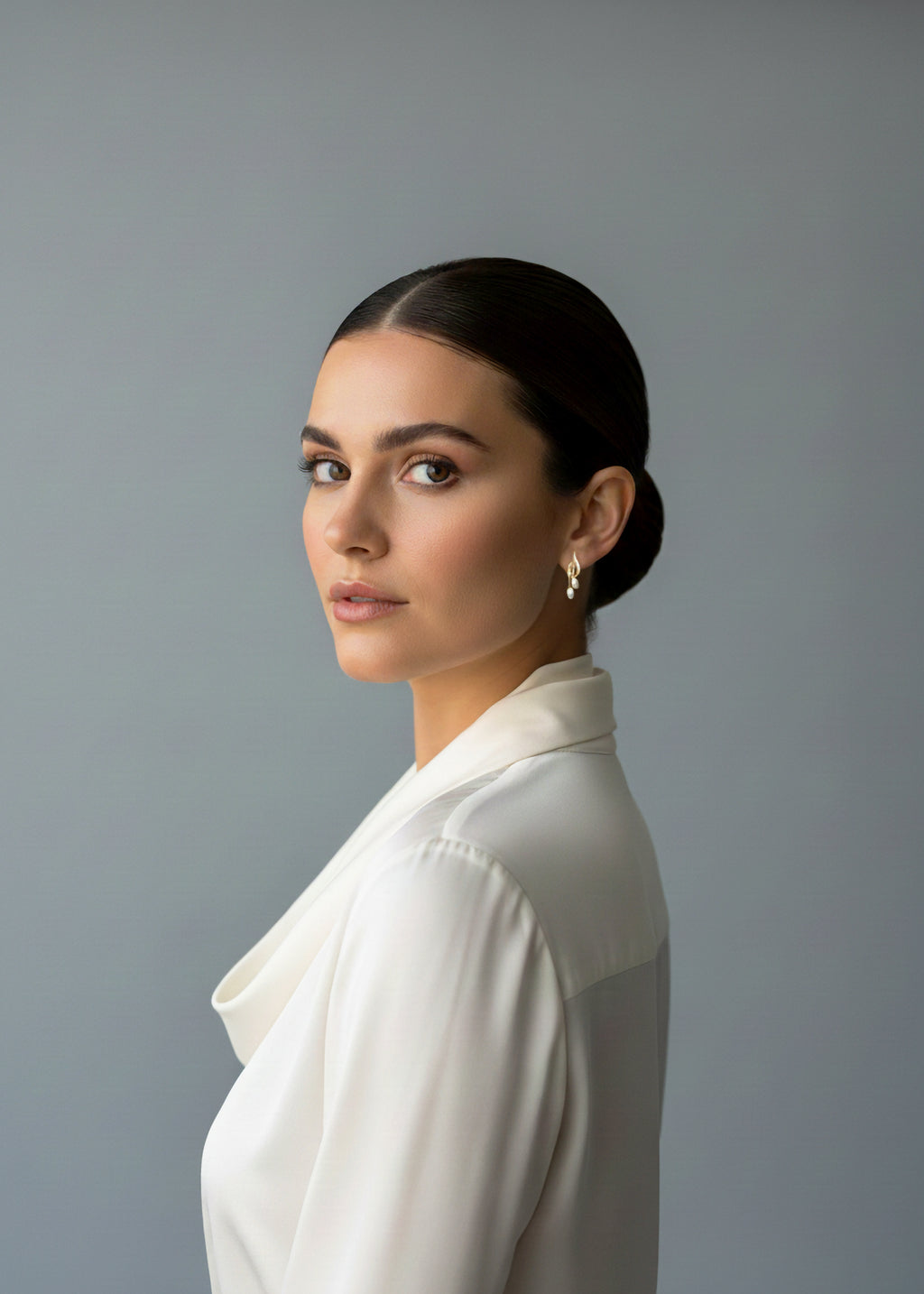 A professional studio portrait of a young woman with dark, sleek hair pulled back into a low bun. She is looking over her shoulder with a calm expression. She wears a white, silk-like blouse with a soft collar and a delicate gold earring with a small pearl drop. The background is a solid, neutral gray.