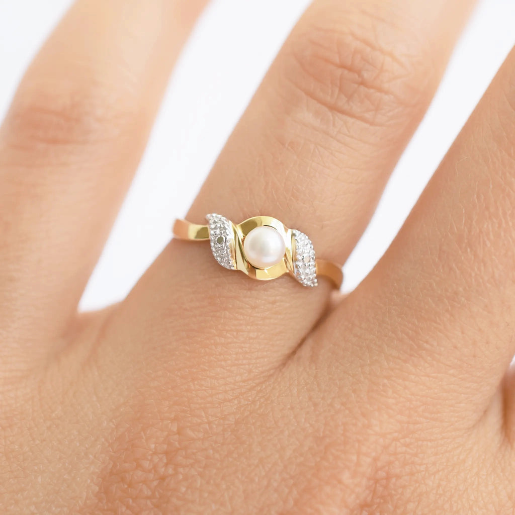 An elegant 18k gold bypass ring featuring a luminous central pearl, with sparkling textured details on each side, shown on a finger.