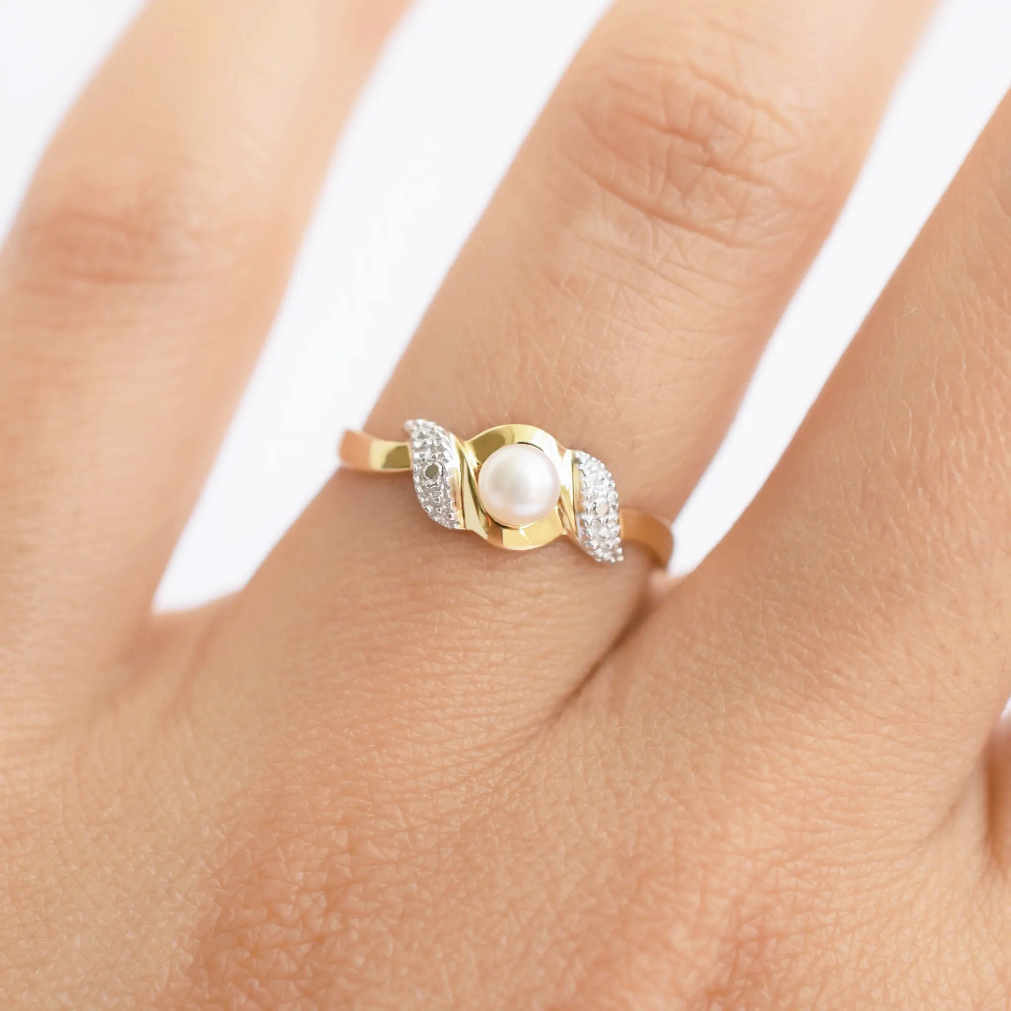 An elegant 18k gold bypass ring featuring a luminous central pearl, with sparkling textured details on each side, shown on a finger.