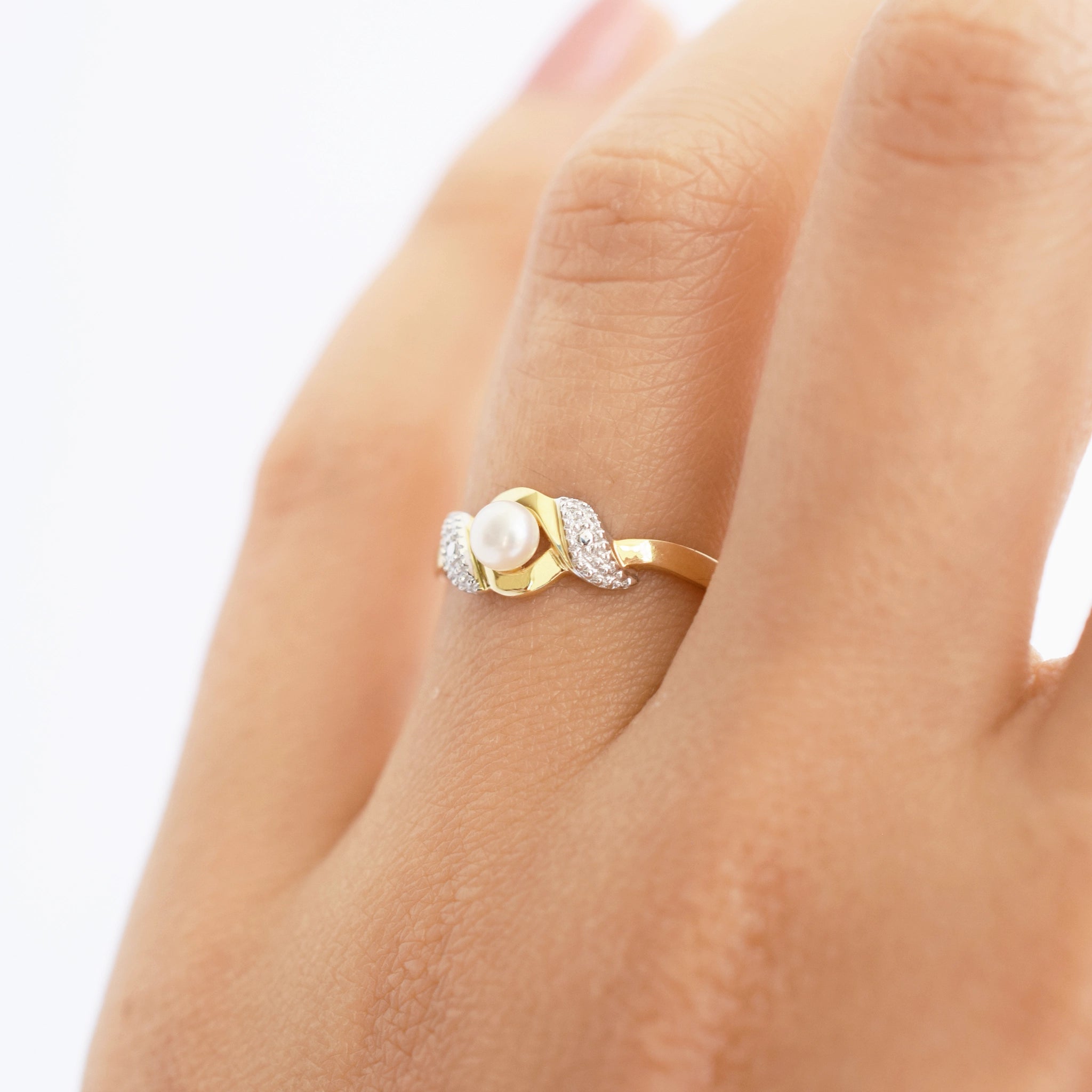 An elegant 18k gold bypass ring featuring a luminous central pearl, with sparkling textured details on each side, shown on a finger.