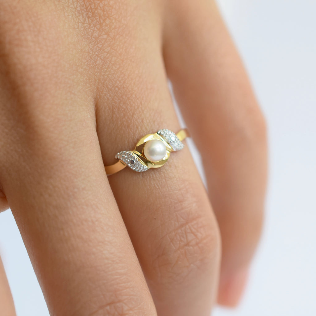 An elegant 18k gold bypass ring featuring a luminous central pearl, with sparkling textured details on each side, shown on a finger.