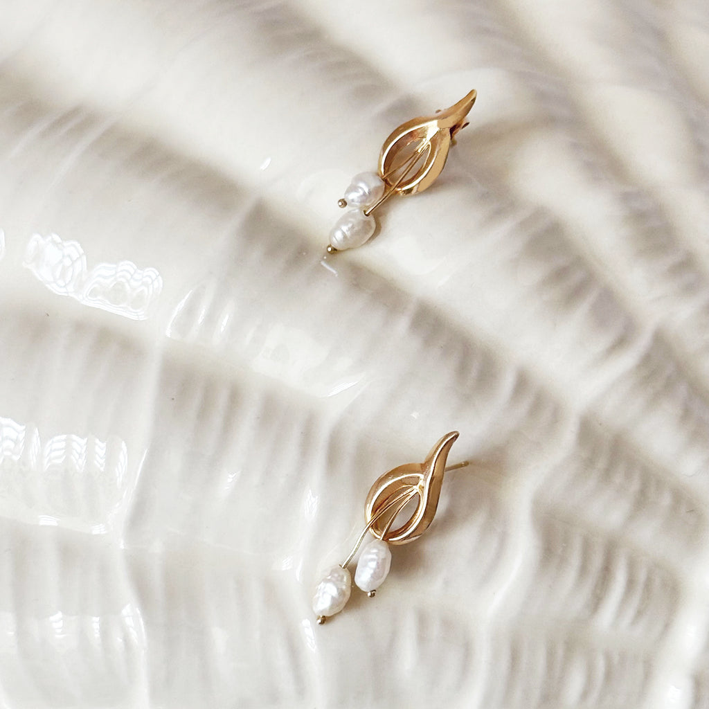 A pair of elegant 14K gold earrings, each featuring a polished, abstract gold top piece with openwork details. From each gold design, two irregularly shaped white baroque pearls dangle at varying lengths. The earrings are displayed side-by-side on a light, subtly textured, off-white surface.