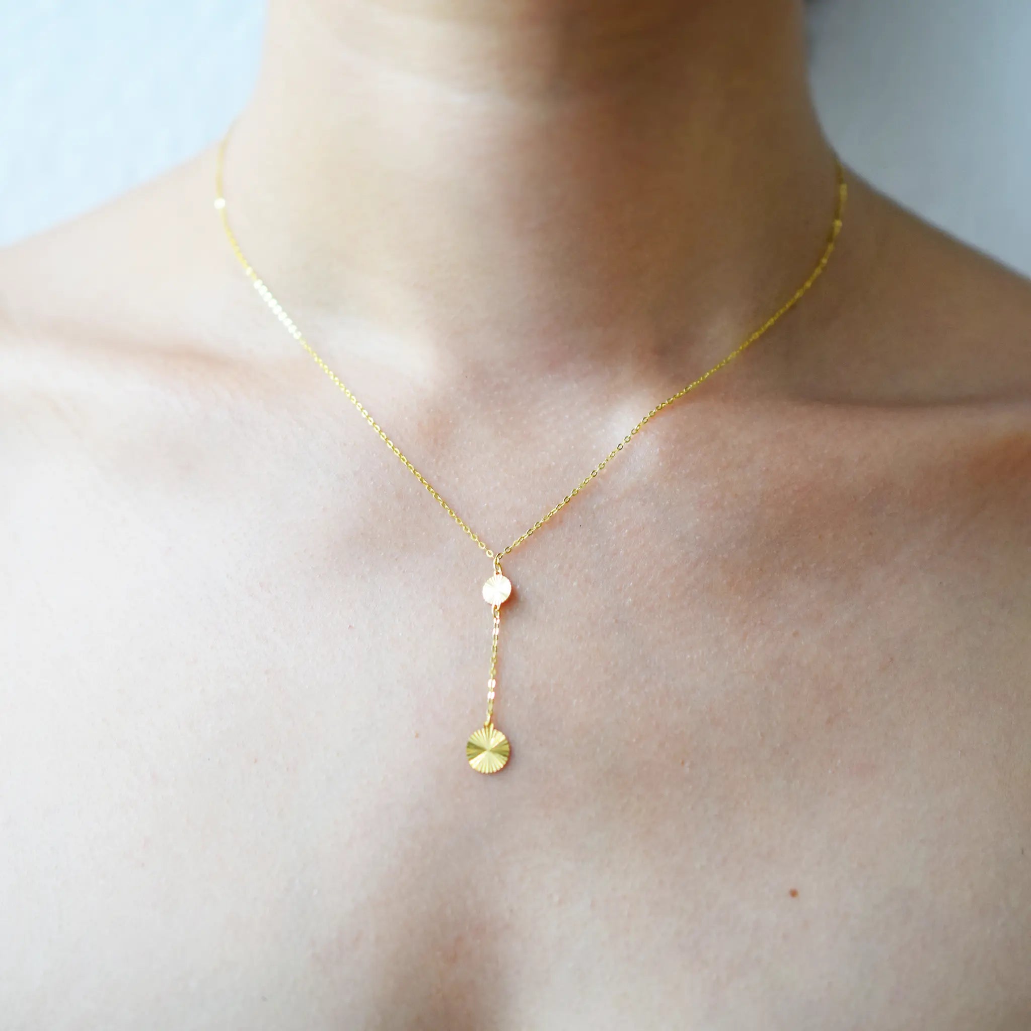 Close-up of a woman wearing a delicate 18k gold lariat necklace. The fine chain features a Y-drop with two small, textured gold discs resting on her décolletage.