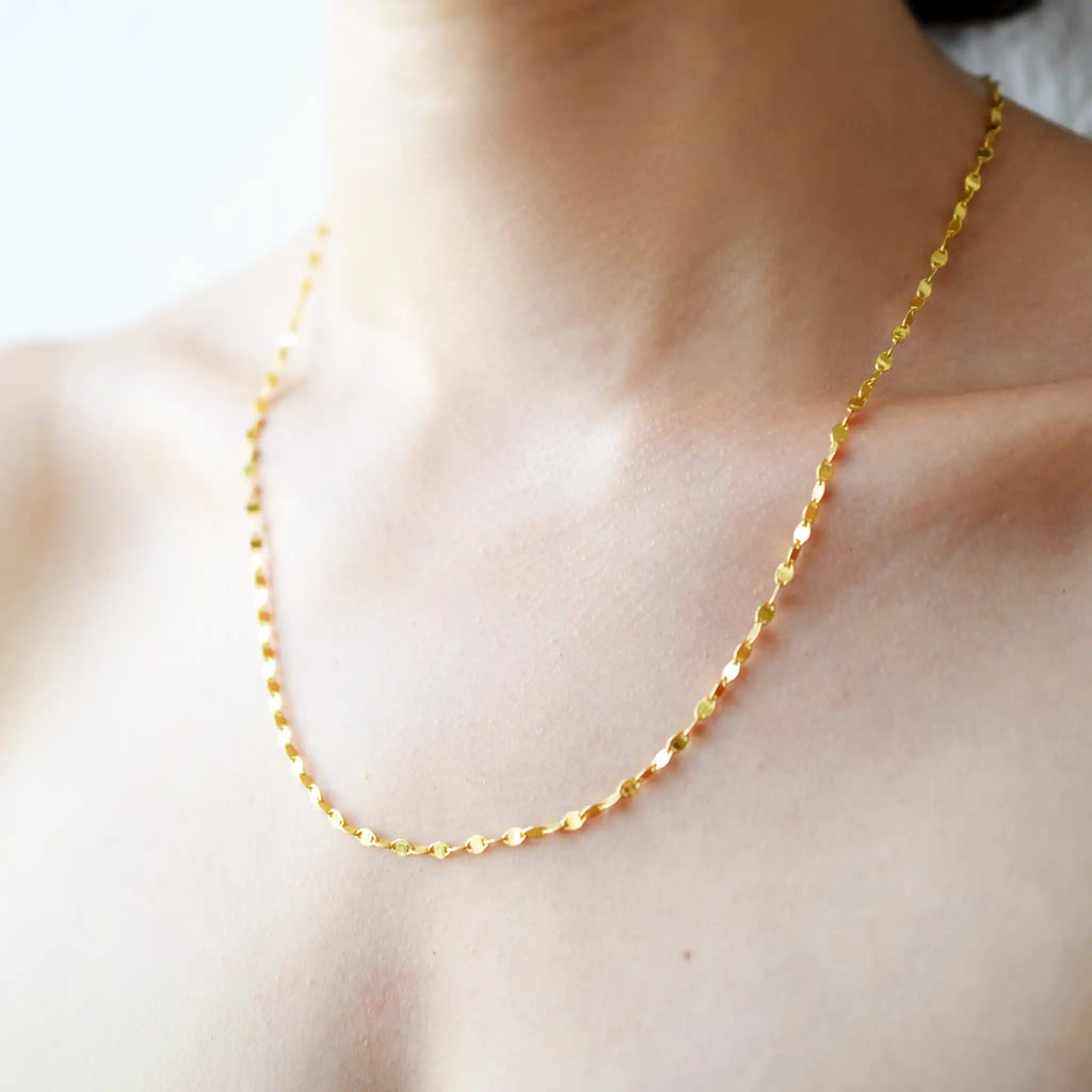 Delicate 21 Karat Yellow Gold necklace featuring a shimmering flat link chain that creates a subtle twisted effect when worn, visible on a model's neck.