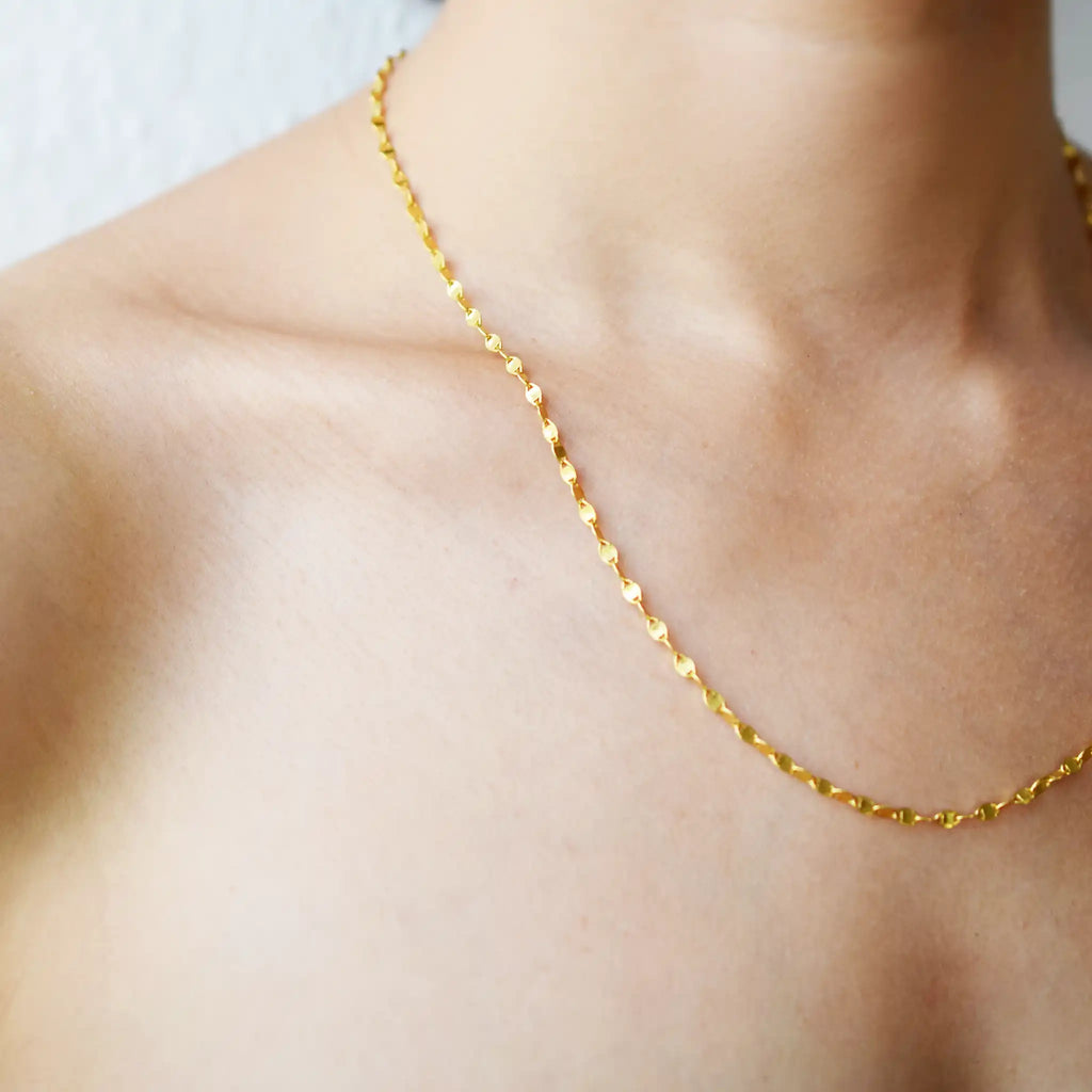 Delicate 21 Karat Yellow Gold necklace featuring a shimmering flat link chain that creates a subtle twisted effect when worn, visible on a model's neck.