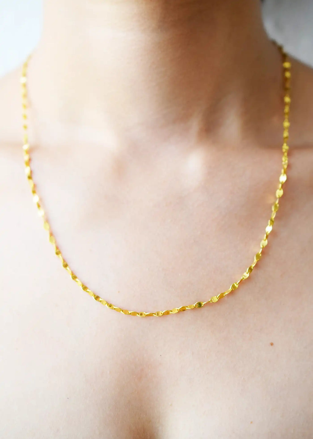 Delicate 21 Karat Yellow Gold necklace featuring a shimmering flat link chain that creates a subtle twisted effect when worn, visible on a model's neck.