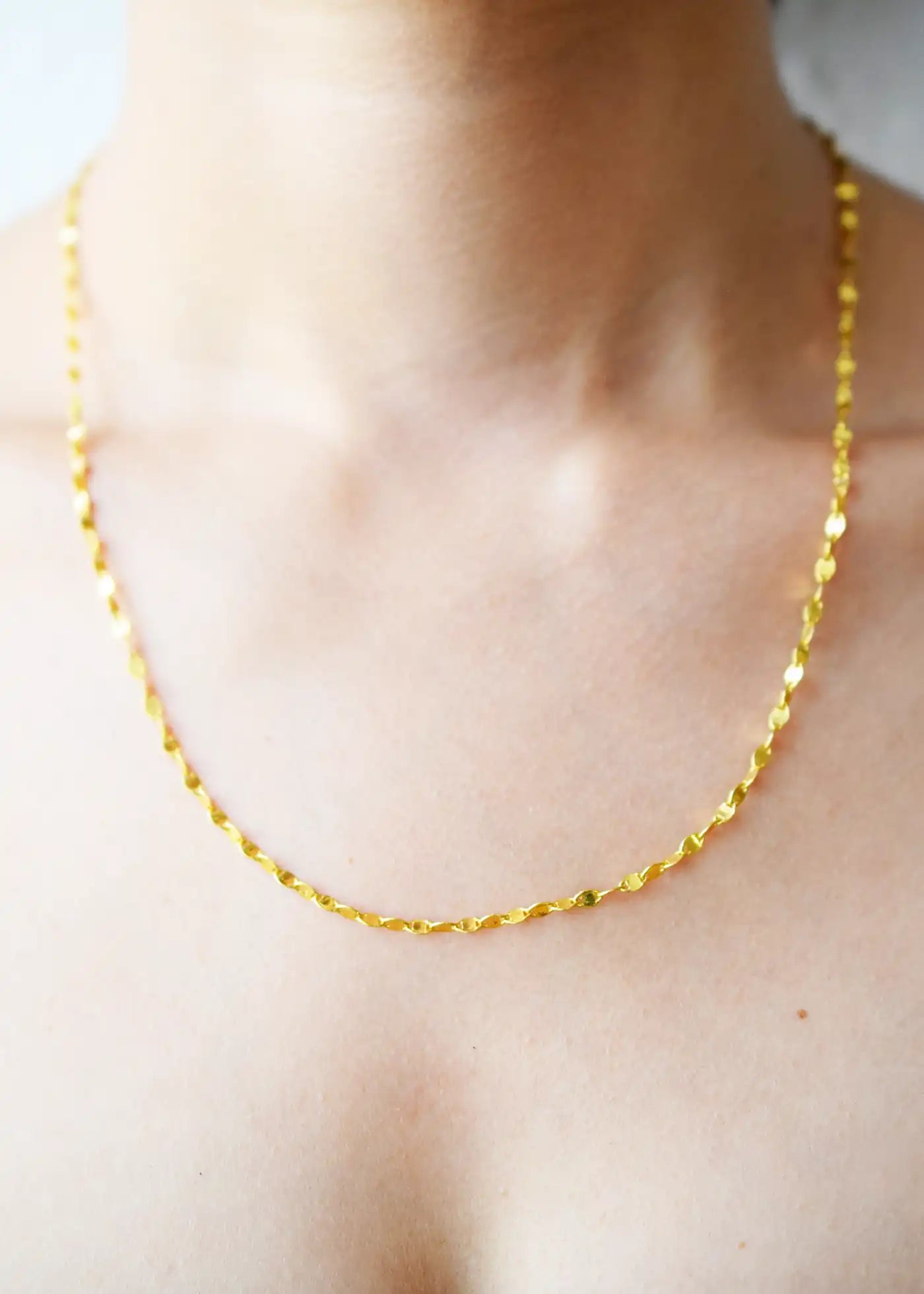 Delicate 21 Karat Yellow Gold necklace featuring a shimmering flat link chain that creates a subtle twisted effect when worn, visible on a model's neck.