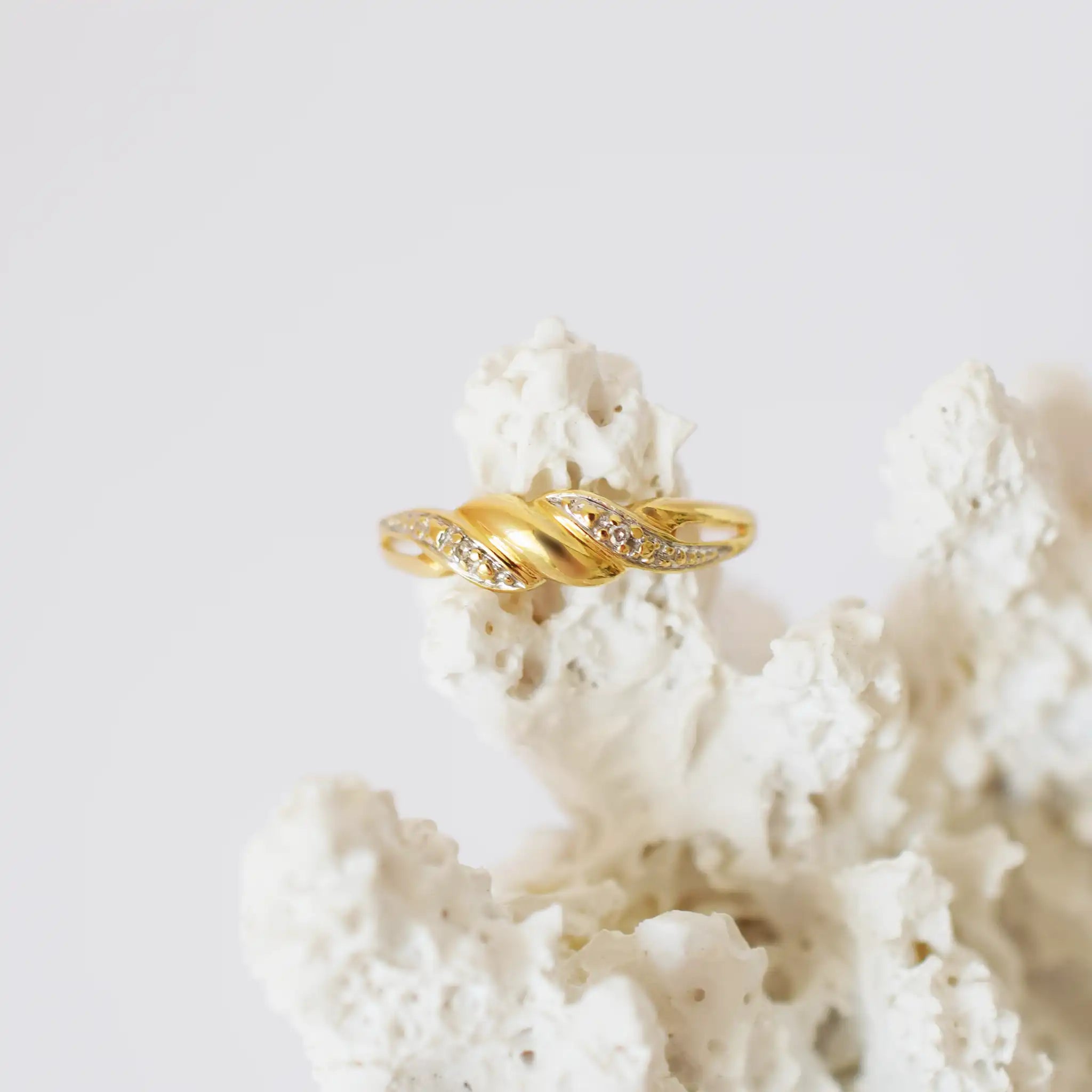 A close-up of an elegant 18K yellow gold ring featuring a twisted band design. The ring incorporates sections of white gold or a silver-toned metal, intricately set with multiple brilliant-cut diamonds along its curves. The ring is displayed on a textured, white, coral-like surface, creating a clean backdrop that highlights the jewelry's intricate details and sparkle.