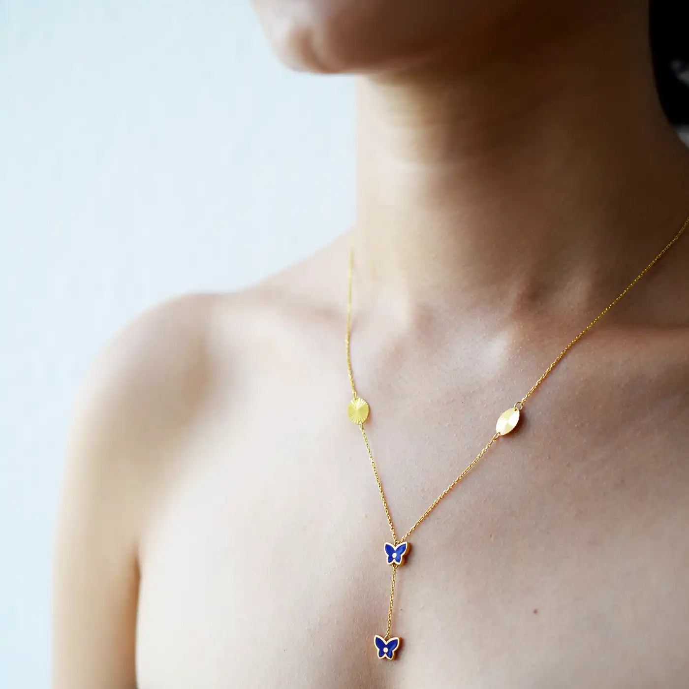 Vibrant 21 Karat Yellow Gold Y-necklace featuring two vivid blue enamel butterfly charms and two grooved gold disc stations along the chain, worn on a model's neck.